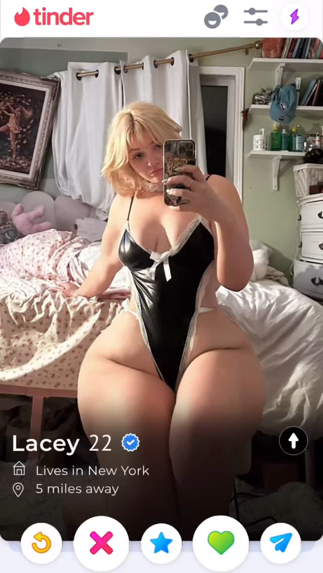 A selfie taken on a dating app interface. The woman is sitting on a bed and holding the phone with her right hand. She is looking at the camera, her expression is neutral. She is wearing black lingerie. The background is a bedroom with curtains, a painting, and items on shelves.