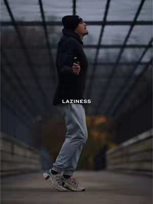 A man wearing a black beanie and jacket is jumping rope on a bridge. The background is blurred, with muted tones and shadows. The text overlay 'LAZINESS' is placed below the figure.