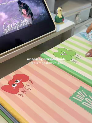 The image showcases a collection of notebooks arranged on a surface. The focus lies on vibrant stationery items. Each notebook features unique patterns, including stripes, and playful graphic elements, such as cartoonish characters. The color palette is dominated by pastel shades, particularly pink, green, and yellow, which impart a sense of cheerfulness and creativity. At the top of the image, a digital tablet displays a video, suggesting the context of the content might be related to lifestyle or unboxing videos. The overall layout of the image creates visually appealing aesthetics, evoking an atmosphere associated with studying or stationery enthusiasm.