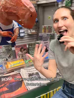 A selfie-style image taken inside a store, featuring a woman with wide eyes and an open mouth, holding a small microphone. She is gesturing with one hand. To the left, a large, orange, dinosaur head prop is visible. In the background are shelves of magazines and other items.