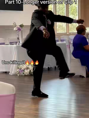 A man in a black suit and hat dances energetically in a room with tables set for an event. He is captured mid-dance, with one leg raised and arms extended. The background includes windows, a white table, and another person seated.