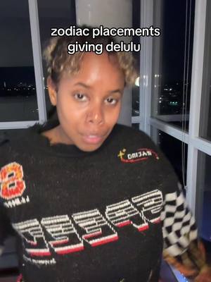 A woman with short hair stands in front of a window at night. The background view is of city lights blurring into the darkness. She is wearing a dark sweater a graphic design. Text overlayed on the image reads \"zodiac placements giving delulu\".