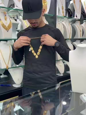 A man wearing a black cap and long-sleeved shirt holds up a gold necklace. The background shows displays with jewelry.