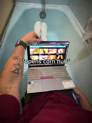 A first-person perspective shot of a person in a bathtub, with feet in white socks visible. They are holding an open laptop showing content on the screen. The hook is overlaid on the image, and has the words 'opens corn hub'. The person's arm and a tattoo are in the bottom left.
