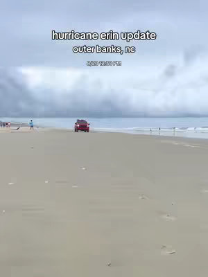 The image shows a beach scene during what appears to be a hurricane. The sky is overcast with heavy clouds. A red vehicle is driving on the beach toward the center of the frame. People are visible in the distance, some near the water's edge, suggesting activity despite the weather. Dunes and the ocean form the background.
