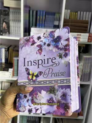 A hand holds an open Bible with a purple floral design on its cover, set against a backdrop of bookshelves filled with books. The Bible's title, "Inspire Praise," is prominently displayed. The overall composition conveys a theme of faith and creativity.