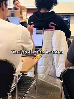 A view of a classroom setting with three people in view. Focus is on the person in front who is working on a laptop on a wooden desk. The person is in a white shirt. Behind the person is another person in a black hoodie with a large design on the back. In the background, a person can be seen with a coffee cup looking at something on the laptop.