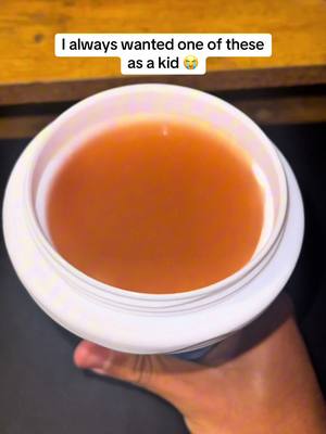 A hand holds a white container filled with a translucent, brownish liquid against a dark background. The container is held at an angle with the liquid visible in the center, and a text overlay floats at the top of the image also.