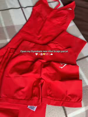 A close-up shot of red Gymshark workout clothes laid out on a patterned surface. The clothes include a tank top and shorts or leggings. Text overlayed on the image says "Open my Gymshark new Vital Sculpt parcel", with a few small emojis.
