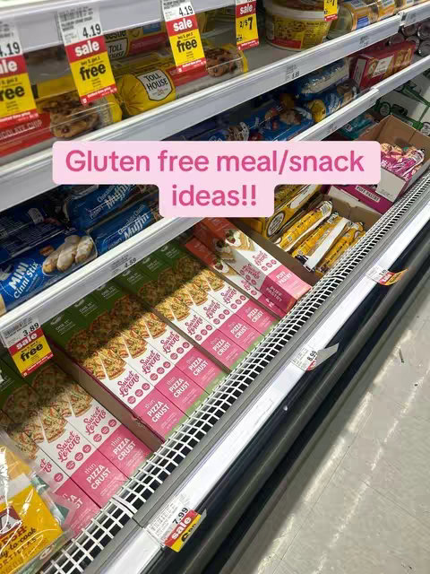 The image shows the inside of a grocery store, with shelves packed with various food items. The camera angle is from a slightly low perspective, looking up at the shelves. There is a light pink rectangular text overlay that reads ‘Gluten free meal/snack ideas!!’ The products on the shelves are organized and labeled, including items like cookies, pizza crusts, and other snacks.