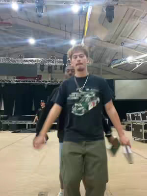 A man with light-colored hair and a mustache is the central figure, positioned with arms extended, holding a beverage container in one hand. He wears a black t-shirt with a car graphic, olive-green shorts, and a silver chain. Behind him, two other individuals are partially visible in the background, in a large building with stage lights and equipment. The image has a candid, informal feel, with an open space and a focus on the central male figure.