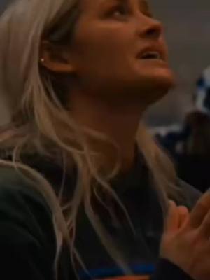 A woman with blonde hair looks upwards with an emotional expression, possibly crying. Her hands are clasped in front of her, and her face is lit with warm lighting.