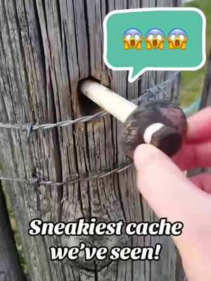 A close-up shot of a weathered wooden fence post with a small cylindrical container inserted into a drilled hole. The container appears to be a geocache. A piece of barbed wire runs horizontally across the post. The image is overlaid with emojis and text. A hand with a red fingernail is pointing at the cache.