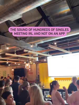 A crowded indoor scene with many people, likely at a social event. The background features industrial-style ceilings, hanging lights, and a bar area. A text overlay in a pink bubble indicates the purpose of the event.