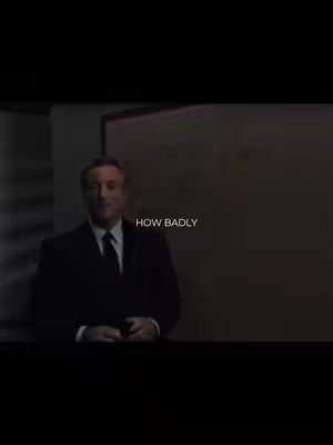 The image is a medium shot of a man in a suit, seated in what appears to be an office or meeting room. The lighting is dim, creating a somber atmosphere. Text overlayed in the image reads, "HOW BADLY". The focus is on the man, who appears to be in mid-speech, giving a motivational feel.