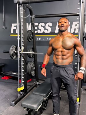 A muscular Black man stands shirtless in a gym, posing in front of a weight machine. To his right, a weight rack, and other gym equipment are visible, creating a gym setting. The man has a tattoo on his chest, and the background shows the gym's logo.