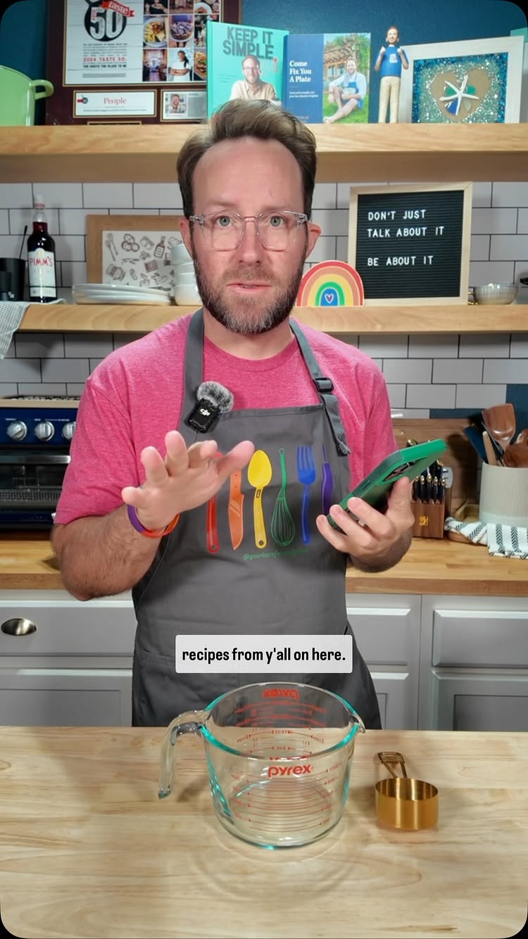 The image features a man in a kitchen setting. He is wearing glasses, a red t-shirt, and a grey apron. He is holding a green phone in his right hand. The background includes shelves with cookbooks and decor. The overlay text says: recipes from y'all on here.