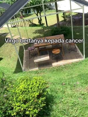 An outdoor shot captured from a high angle. It features a gazebo-like structure with white framing. Inside, there's a wooden table surrounded by stools. The scene is set on a platform-like surface, with lush green grass surrounding it, interspersed with a small bush in the foreground. Text overlay stating "Virgo bertanya kepada cancer" is added at the bottom.