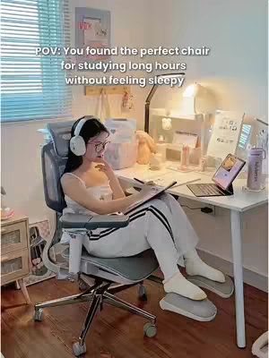A well-lit room with a desk and an ergonomic chair. A woman with eyeglasses and headphones sits in the chair, looking at a tablet. The chair has a headrest and armrests. A window and blinds are visible in the background. The image has a bright, clean aesthetic.