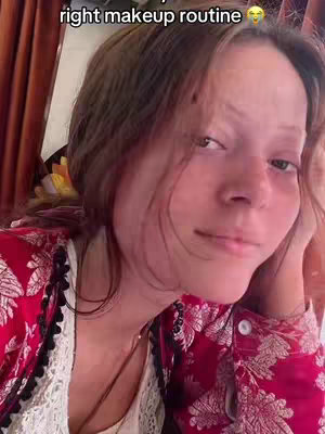 A woman with light skin and brown hair looks toward the camera. She wears a red and white patterned top with a lace neckline and dark necklace. Her right hand is held up against her cheek. The background includes neutral tones.