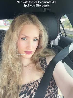 A woman with long blonde hair is sitting in a car, looking at the camera. She has makeup on, including eyeliner and lipstick. She is wearing a leopard-print top and a seatbelt is across her chest. The background shows the interior of the car and a view of an exterior scene.
