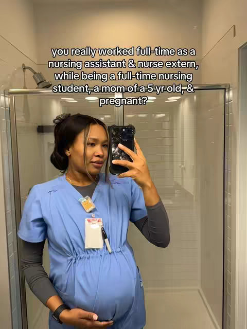 A woman takes a selfie in a bathroom, holding her phone up to the mirror. She is pregnant and wearing a light blue medical scrub top. The background is a clean, white bathroom with a glass shower door visible. Overlay text is included above.
