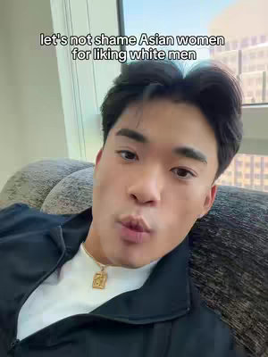 A young Asian man with short black hair is centered in the frame, looking directly at the camera with a slight pout. He's wearing a white t-shirt under a black jacket and a gold pendant necklace. The background features a muted interior scene, with a glimpse of a window.