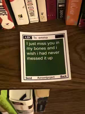 A close-up shot of a small square card resting on a wooden surface in front of a shelf of books. The card has a green background with text in white, and borders with details related to who the message is addressed to and the sender. The text says, "I just miss you in my bones and I wish I had never messed it up".