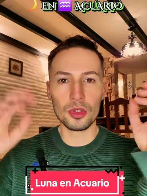 A man is the central focus, framed from the chest up against a domestic setting. The background features a light-colored, textured wall adorned with a small framed picture and a ceiling with visible wooden beams. Overlaying the image are text elements, including a horizontal sign at the bottom with the words \"Luna en Acuario\" in white font against a red background, and another text element at the top with text in a green frame.