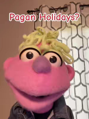 The image features a puppet with a large pink face, large black eyes, a purple nose, and green hair. The puppet is wearing a denim jacket, and behind it is a patterned curtain.