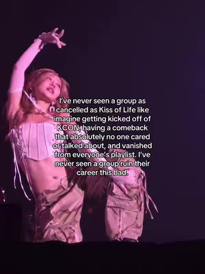 A woman with light skin and light-colored hair is on stage in a show setting. She is in the middle of the frame with her left arm raised overheard and index and middle finger up in a peace sign, and her right arm is bent with her hand in a fist. She is wearing a light pink, small top and tan/brown pants. The background is dark but has some lighting to it.