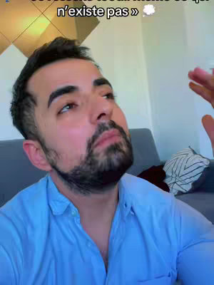 A selfie-style shot features a man looking upwards, with his face framed in a close-up. He has a dark beard and is wearing a light blue button-down shirt. The background suggests an indoor setting.