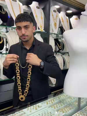 A man in a black suit stands in a jewelry store, holding a large gold necklace. The background is filled with displays of gold jewelry. The man is looking at the camera.