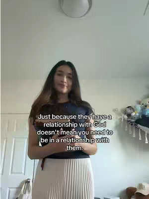 A woman is standing in front of a plain wall. Her hair is dark, and she is wearing a navy blue top and a white pleated skirt. Text overlayed on the image reads: "Just because they have a relationship with God doesn't mean you need to be in a relationship with them."