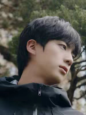 The cover image is a medium shot of a young man, likely Jin, looking off to the side with a thoughtful or contemplative expression. The background is slightly blurred, showing trees and foliage, suggesting an outdoor setting. The man is wearing a dark jacket, and his dark hair is neatly styled. The lighting appears natural, possibly daylight. The composition focuses on his face and upper body, with the background providing a natural backdrop.