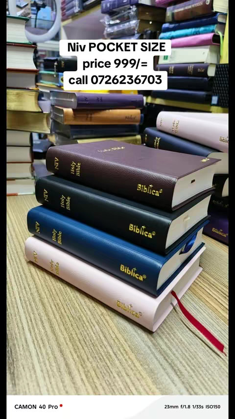 The image showcases a vertical display of stacked hardcover Bibles. The Bibles are primarily leather-bound in various colors, including dark brown, black, blue, and light pink. The titles 'Holy Bible' and 'Biblica' are prominently displayed on the spines and covers in gold lettering. An overlay at the top contains white text advertising 'Niv POCKET SIZE,' with the price and a phone number (0726236703) for purchasing. The background appears to be a display shelf potentially in a bookstore, with other books visible.