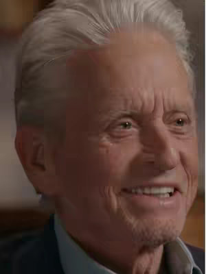 A close-up shot of Michael Douglas, focusing on his face and upper chest. He is looking slightly to the right with a subtle smile. His silver hair is neatly styled, and the lighting emphasizes the details of his face; wrinkles and creases are prominent. He's wearing a dark blue blazer with a light blue shirt visible underneath.