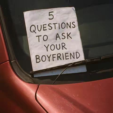 A square image showing a handwritten sign stuck to what appears to be a car window. The sign, made of plain white paper, has the text '5 QUESTIONS TO ASK YOUR BOYFRIEND' in large, uneven, black handwriting. The background is a blurred view through the car windshield.