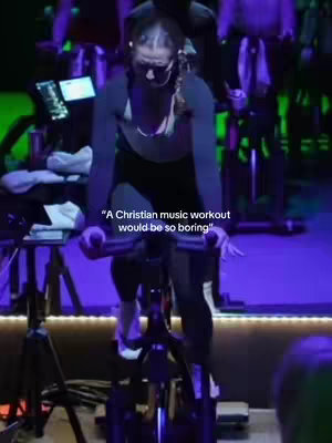 The image shows a woman riding a stationary bike in a cycling studio. She is wearing workout attire, including a black top and leggings. The studio is illuminated with purple and green lights, and other cyclists are visible in the background. Text overlay on the image reads, “A Christian music workout would be so boring”.