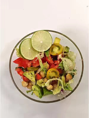 A clear glass bowl filled with a colorful salad. The salad features various ingredients including sliced tomatoes, avocado, lettuce, and chickpeas. Two lime slices are arranged on top. The bowl is set against a plain white background.