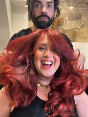 A woman with red hair and red lipstick is smiling widely, with a man behind her, presumably the hairstylist, with a beard and dark hair. The woman is wearing a black tank top and necklace. The background is blurred, suggestive of a salon setting.