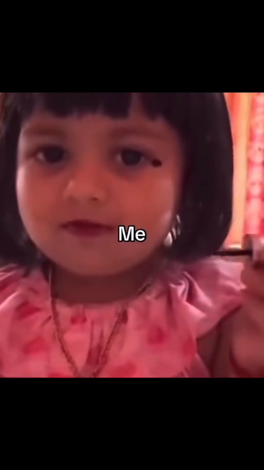 A young girl with a blunt black-haired bob is holding a makeup brush towards her left eye. The background is a blurred pink and red.