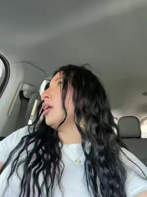 A young woman with long, dark hair is seated in a car, looking out the side window. She wears a white t-shirt and a delicate necklace, and the interior of the car and the passing scenery outside are visible. The lighting appears to be natural and ambient.