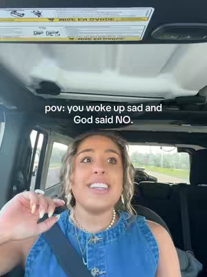 A woman with curly blonde hair is sitting inside a car, likely a Jeep, looking toward the camera, smiling. The image is shot from a first-person perspective. The text above the woman reads: 'pov: you woke up sad and God said NO.' The woman is wearing a denim top and a necklace. The background is blurred, suggesting movement or a shallow depth of field. The overall tone is lighthearted and conversational, likely targeting a relatable experience.