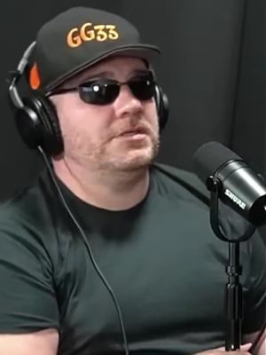 A man wearing a black cap with 'GG33' written on it, sunglasses, and a headset. A microphone is positioned on the right side. The background is dark.
