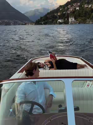 A video cover image shows a woman reclining on the front of a boat, reading a book. A man is driving the boat. The scene is set on a lake, with mountains and buildings in the background.