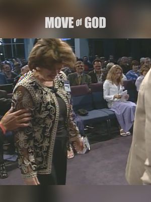 The image shows a woman in a church setting, possibly receiving a blessing. She is the central figure, dressed in a dark outfit with an ornate, sheer, black and white patterned overcoat. Her head is bowed, and she appears to be in a moment of emotion or spiritual experience. Two hands are visible, possibly touching her arm, suggesting support or prayer. In the background, a seated audience observes the scene. The lighting is soft, with a focus on the woman, and the overall composition suggests a moment of religious significance.