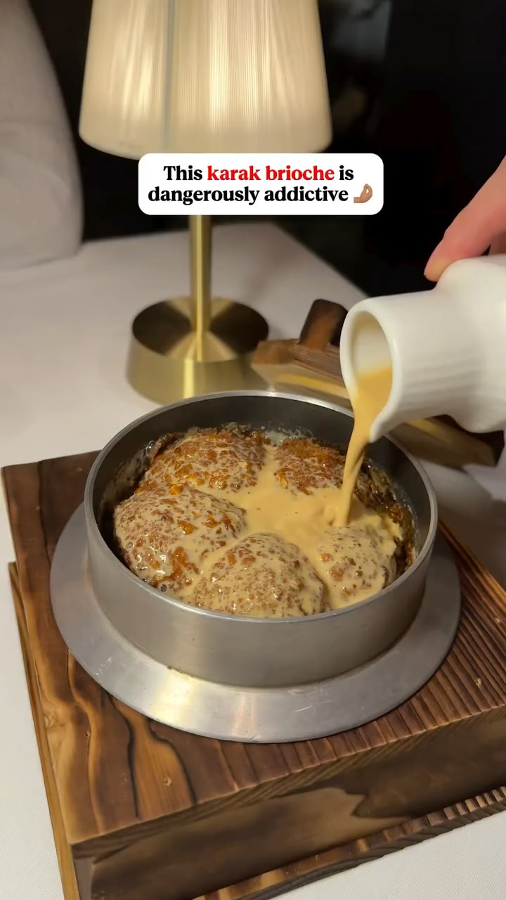 A close-up shot of a dish with a dark brown wooden base. The dish, a metallic vessel with a handle, contains several pieces of golden-brown, possibly baked, items that are partially covered in a light, creamy sauce. A hand is seen pouring more of the sauce from a white, cylindrical pitcher. In the background, a warm-toned lamp provides soft lighting, and a white surface likely represents a table or countertop. The composition highlights the dish, emphasizing the food and the pouring action.