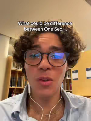 A close-up shot of a young man with curly hair, wearing glasses and a light blue button-down shirt. He is looking directly at the camera and speaking. The background includes a bookcase and yellow wall.