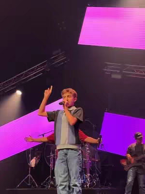 A young man on a stage with a microphone, singing and gesturing upwards. He's wearing a short-sleeved shirt and jeans. Behind him, there are other musicians and large, bright purple panels overhead. The stage is well-lit, and the background is dark.